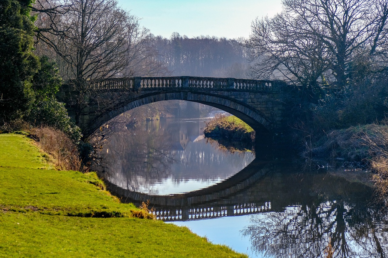 A stone bridge over a body of water surrounded by green grass and bare trees, with a building in the distance and the scene captured in the water's reflection